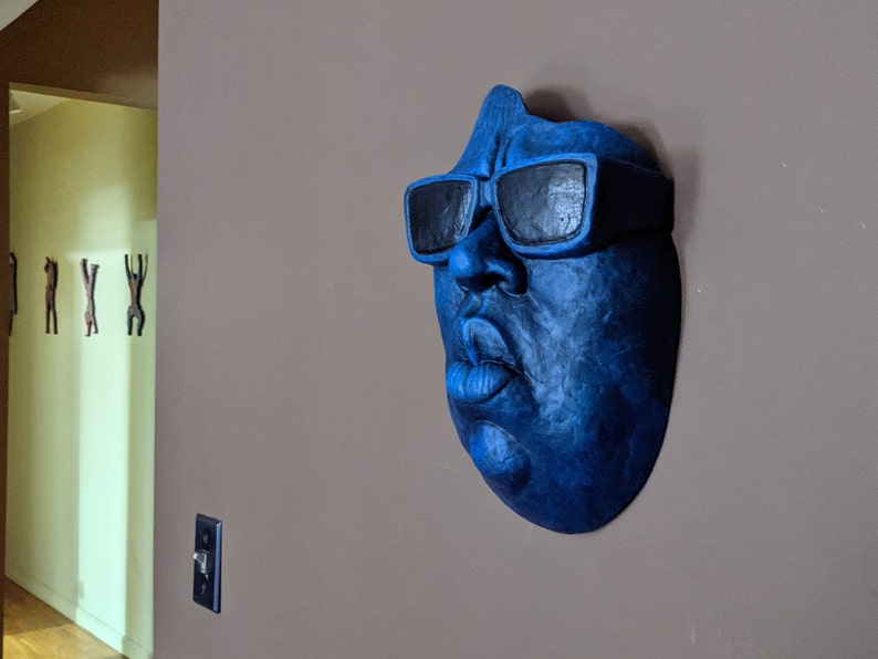 May include: A blue wall sculpture of a face wearing black sunglasses. The sculpture is mounted on a neutral-colored wall. The face has a prominent nose and lips. The sculpture is a unique piece of wall art.