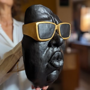 May include: A black sculpture of a face wearing gold-rimmed sunglasses. The sculpture has a textured surface and detailed facial features, including a prominent nose and full lips. The sunglasses have black lenses.