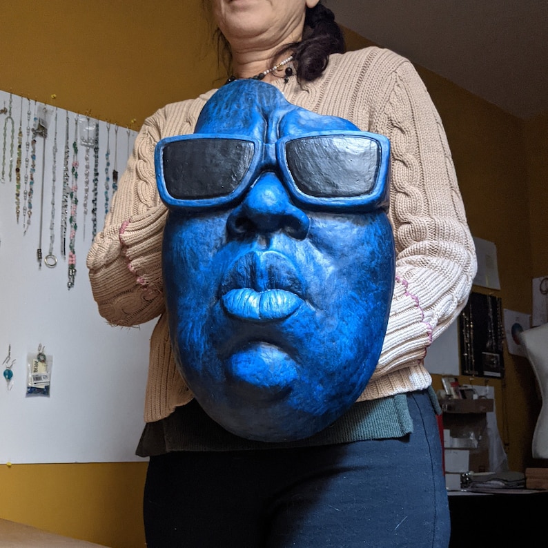 May include: A large, blue face mask with black sunglasses. The mask has a textured surface and is held by a person wearing a tan sweater and black pants. The mask's features include a nose, lips, and eyebrows.