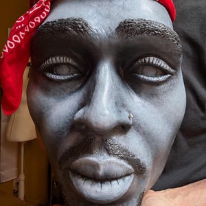 May include: A sculpted bust of a man with a red bandana. The face is painted in shades of gray and black, with detailed features including a nose ring and a goatee. The bandana is red with a white pattern.