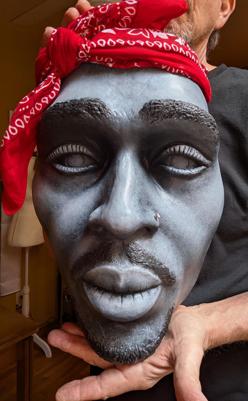 May include: A sculpted bust of a man with a red bandana. The face is painted in shades of gray and black, with detailed features including a nose ring and a goatee. The bandana is red with a white pattern.