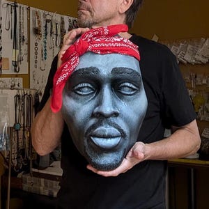 May include: A gray sculpture of a face with a red bandana. The sculpture has a goatee and a nose ring. The person holding the sculpture is wearing a black shirt.