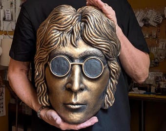 May include: A bronze-colored sculpture of a face with round glasses and long hair. The sculpture is held up by a person. The face has a detailed depiction of the eyes, nose, and mouth. The sculpture is a bust of a famous person.