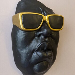 May include: A black sculpture of a face wearing gold-rimmed sunglasses. The sculpture has a matte finish and is mounted on a light pink wall. The sunglasses have black lenses and a thick gold frame.