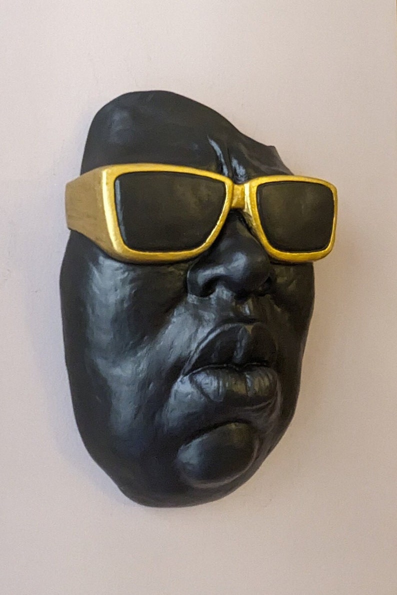 May include: A black sculpture of a face wearing gold-rimmed sunglasses. The sculpture has a matte finish and is mounted on a light pink wall. The sunglasses have black lenses and a thick gold frame.