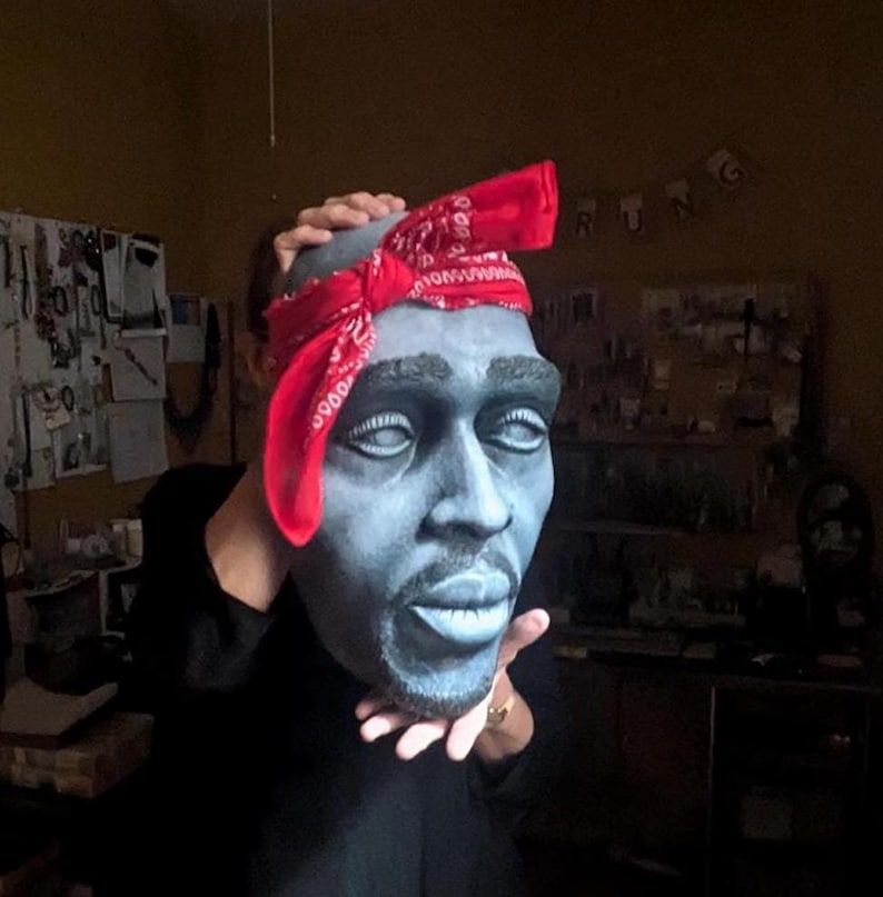 May include: A gray bust sculpture of a man with a red bandana. The sculpture has white eyes and a goatee. The bandana is tied around the head with the ends hanging down. The sculpture is held up by a person.