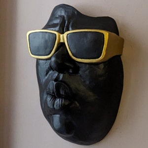 May include: A black sculpture of a face wearing gold-rimmed sunglasses. The face has a prominent nose and pursed lips. The sculpture is mounted on a light-colored wall.