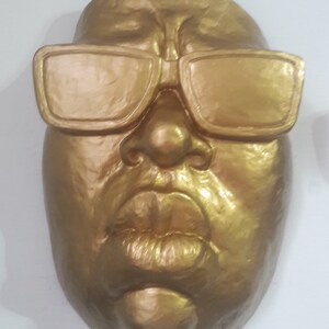 May include: A golden-colored face sculpture with sunglasses. The sculpture features a detailed nose, full lips, and a smooth, reflective surface. The sunglasses are rectangular and also golden.