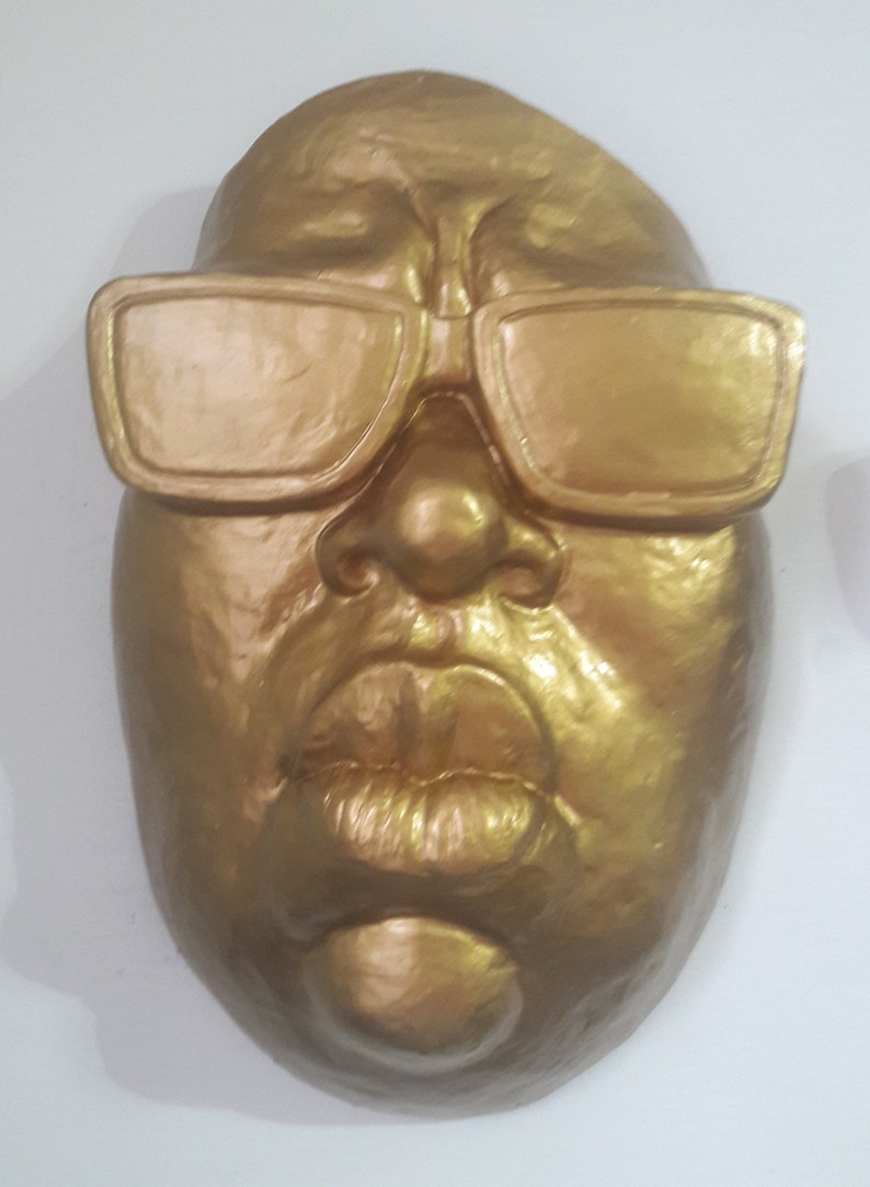May include: A golden-colored face sculpture with sunglasses. The sculpture features a detailed nose, full lips, and a smooth, reflective surface. The sunglasses are rectangular and also golden.