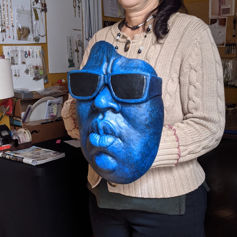 May include: A blue face mask with black sunglasses. The mask has a textured surface and detailed facial features, including lips and a nose. The mask is held in front of a person.