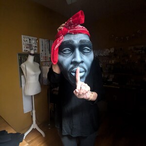 May include: A large, stylized mask of a man's face, painted in shades of blue and gray, with a red bandana. The person is wearing a black shirt and pants, with a finger held up to the lips in a 'shush' gesture.