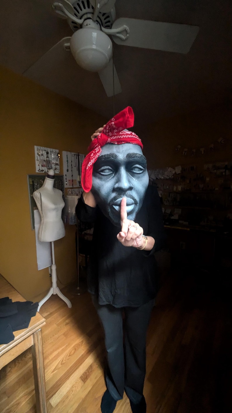 May include: A large, stylized mask of a man's face, painted in shades of blue and gray, with a red bandana. The person is wearing a black shirt and pants, with a finger held up to the lips in a 'shush' gesture.