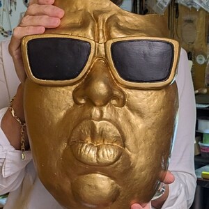 May include: A gold-painted mask with black sunglasses. The mask has a detailed face with a prominent nose and full lips. The mask is held up by a person wearing a white shirt.