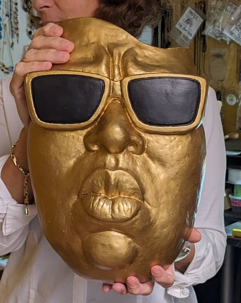 May include: A gold-painted mask with black sunglasses. The mask has a detailed face with a prominent nose and full lips. The mask is held up by a person wearing a white shirt.