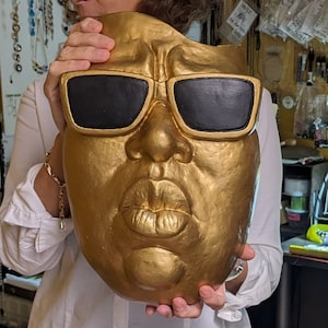 May include: A gold-colored mask with black sunglasses and a large mouth. The mask is held up by a person wearing a white shirt. The mask has a textured surface and is likely made of a solid material.