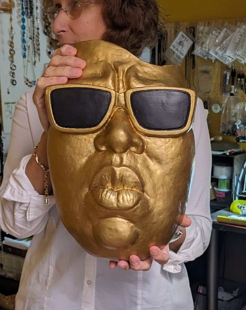 May include: A gold-colored mask with black sunglasses and a large mouth. The mask is held up by a person wearing a white shirt. The mask has a textured surface and is likely made of a solid material.