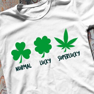Peut inclure: T-shirt blanc avec trois motifs de feuilles vertes : un trèfle à trois feuilles, un trèfle à quatre feuilles et une feuille de cannabis. Les mots "NORMAL", "LUCKY" et "SUPERLUCKY" sont en dessous. Sur une surface en bois.
