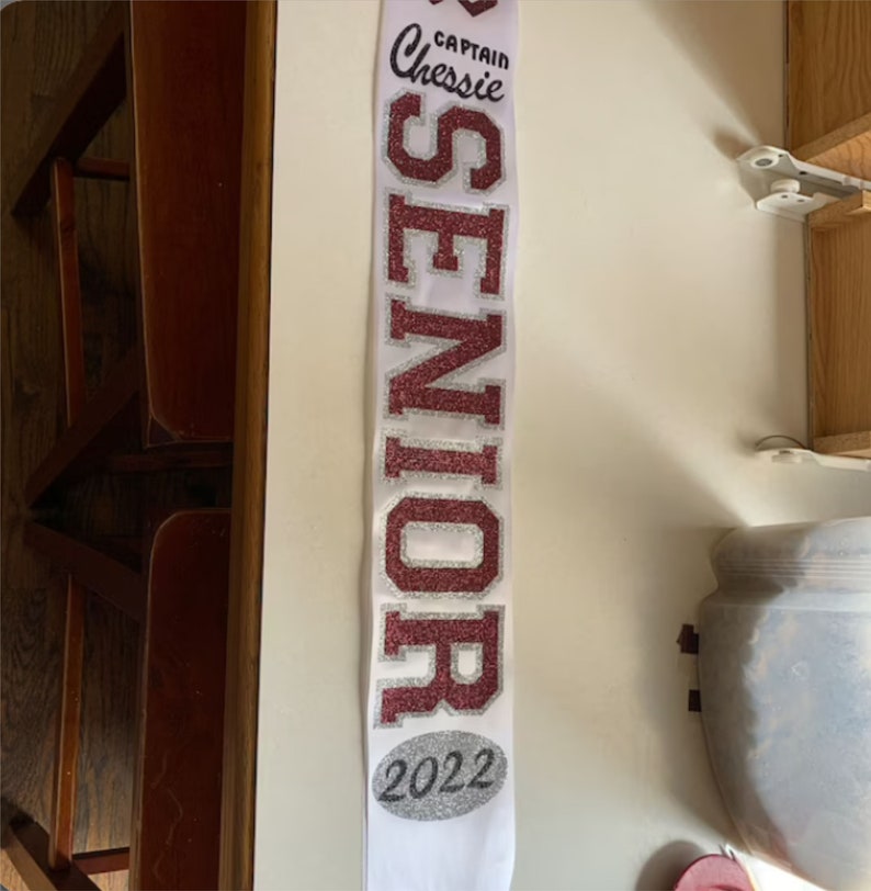 May include: White sash with the word "SENIOR" in large, glittery, burgundy letters. The sash also has the text "CAPTAIN Chessie" and "2022" in smaller, silver glitter letters.