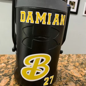 May include: Black insulated water bottle with a yellow and white "DAMIAN" name, a white Under Armour logo, and a yellow and white "B" with the number "27" underneath.