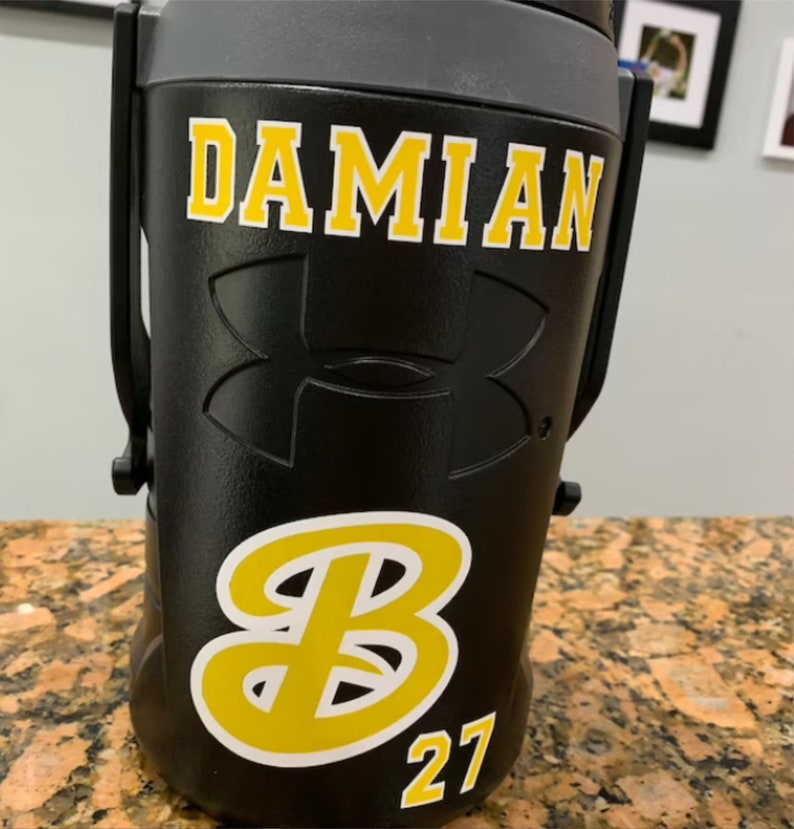 May include: Black insulated water bottle with a yellow and white "DAMIAN" name, a white Under Armour logo, and a yellow and white "B" with the number "27" underneath.
