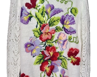 May include: Cream-coloured knitted jumper with a floral pattern. The jumper has a round neck and long sleeves with a decorative openwork design. The floral pattern features red, purple, pink, and yellow flowers with green leaves.