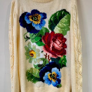 Puede incluir: Un suéter de punto color crema con un diseño floral. Las flores son rojas, azules, amarillas y verdes. El suéter tiene cuello redondo y mangas largas.