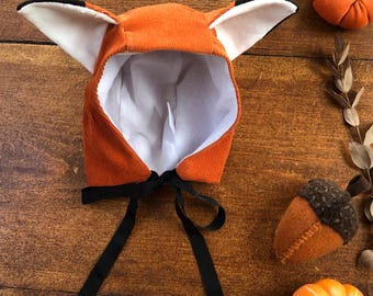 May include: An orange corduroy bonnet featuring two large fox ears with black tips and white inner lining. The bonnet has a white fabric interior lining and two long black ribbon ties attached at the base to secure the head covering under the chin.