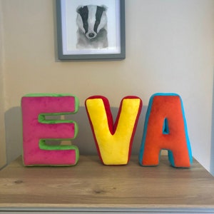 May include: Three colourful plush letters spelling out the name "EVA". The letter E is pink with green trim, the letter V is yellow with red trim, and the letter A is orange with blue trim.
