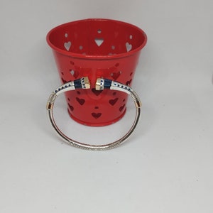 Puede incluir: Un brazalete de plata con detalles azules y dorados. El brazalete está en un recipiente rojo con recortes en forma de corazón.