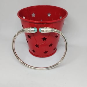 Puede incluir: Un brazalete de plata con detalles turquesa. El brazalete está sobre un recipiente rojo con recortes de estrellas.