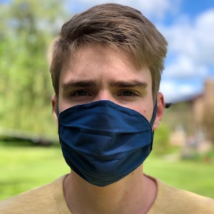 Puede incluir: Una persona joven con una mascarilla facial azul. La mascarilla está hecha de tela y tiene pliegues. La persona lleva una camiseta amarilla.