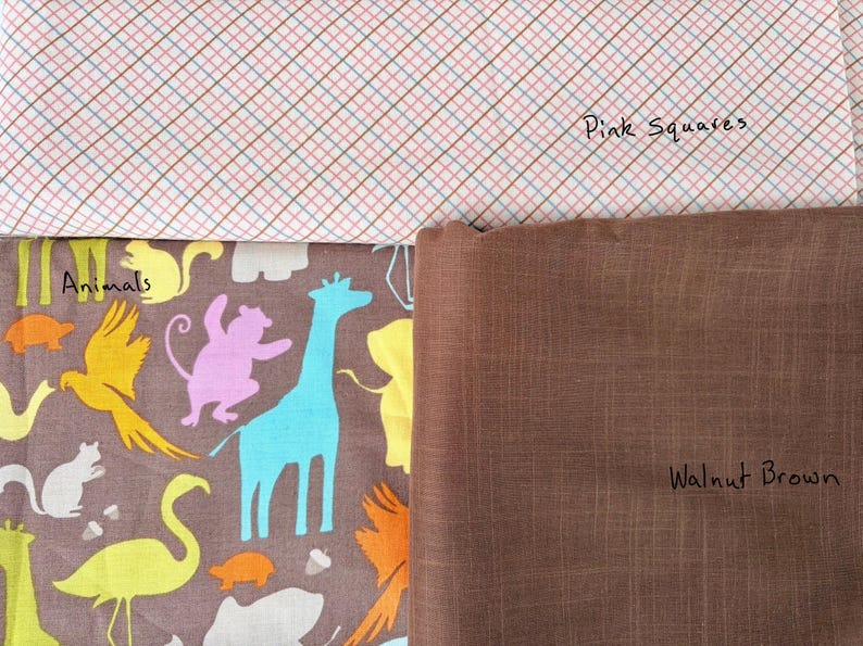 May include: A fabric assortment featuring three distinct patterns. The top fabric displays a pink and white plaid design with the text "Pink Squares." Below, a brown fabric is labeled "Walnut Brown." The bottom fabric showcases colorful animal illustrations.