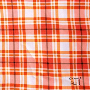 May include: Close-up of a fabric with an orange plaid pattern. The plaid design features a grid of orange, white, and black lines. The words "Orange Plaid" are handwritten in black ink in the lower right corner.
