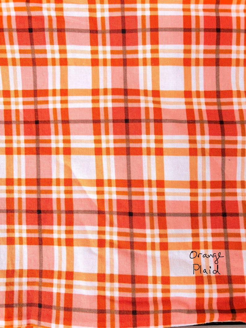 May include: Close-up of a fabric with an orange plaid pattern. The plaid design features a grid of orange, white, and black lines. The words "Orange Plaid" are handwritten in black ink in the lower right corner.