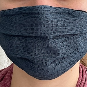 Può includere: Una maschera facciale blu scuro realizzata in tessuto strutturato. La maschera ha un elastico nero su ogni lato.