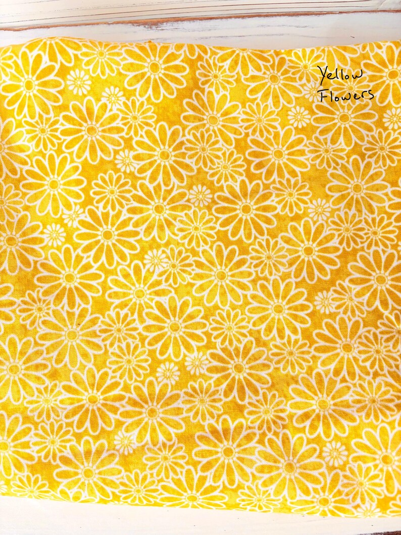 May include: A bright yellow fabric with a repeating pattern of white daisy-like flowers. The words "Yellow Flowers" are handwritten in black ink in the upper right corner. The fabric is likely cotton or a similar material.