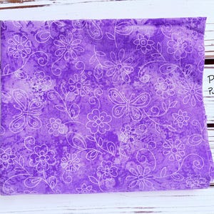 May include: A folded piece of fabric with a purple background and a white floral and butterfly pattern. The fabric has the words "Playful Purple" written on it. The fabric is on a white wooden surface.