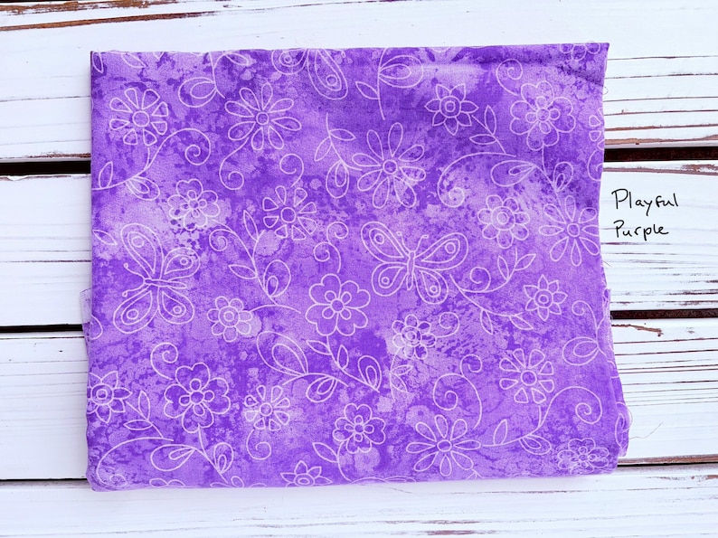 May include: A folded piece of fabric with a purple background and a white floral and butterfly pattern. The fabric has the words "Playful Purple" written on it. The fabric is on a white wooden surface.