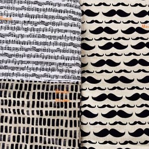 May include: A fabric panel featuring three distinct patterns. One section displays black musical notes on a white background with the word "Music". Another shows a repeating pattern of black mustaches on a beige background with the word "Moustache". The third section has a black and white abstract design with the words "Brush Strokes".