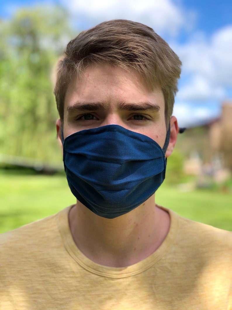 May include: A person wearing a dark blue face mask, covering the nose and mouth, with black ear loops. The person is wearing a yellow t-shirt. The background is a blurred green lawn and blue sky. The image is a close-up.