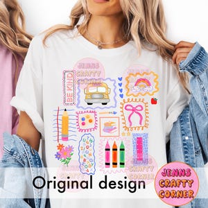 May include: A white t-shirt featuring a colourful school-themed graphic print. The design includes a yellow school bus, a pencil, crayons, a pink bow, a rainbow, books, flowers, and the text "BE KIND" and "JENNS CRAFTY CORNER" in various frames.
