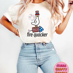 May include: A white t-shirt with a cartoon image of a duck wearing a red, white, and blue hat and riding a firework. The text "fire quacker" is printed below the image.
