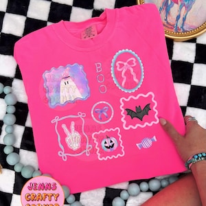 May include: Bright pink sweatshirt with Halloween-themed graphics. The design includes a ghost, bows, a bat, a pumpkin, and candy. The word "BOO" is printed vertically. The text "Jenns Crafty Corner" is also visible.
