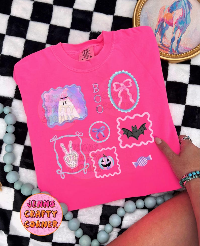 May include: Bright pink sweatshirt with Halloween-themed graphics. The design includes a ghost, bows, a bat, a pumpkin, and candy. The word "BOO" is printed vertically. The text "Jenns Crafty Corner" is also visible.