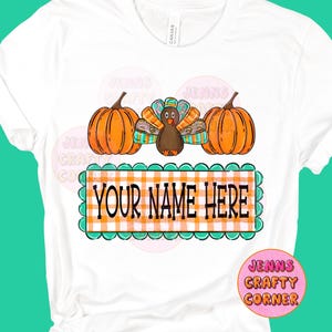 May include: White t-shirt featuring a Thanksgiving design. The design includes two orange pumpkins, a turkey with colorful feathers, and a rectangular sign with the text "YOUR NAME HERE" in black letters on an orange and white checkered background.