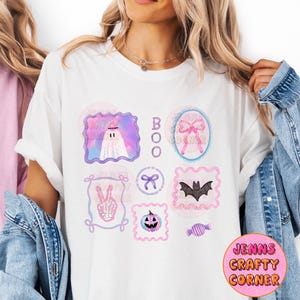 May include: White t-shirt with a colorful Halloween-themed design. The design includes a ghost, a bat, a jack-o'-lantern, and the word "BOO". The shirt also features the text "Jenns Crafty Corner" in a pink circle.