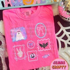 May include: A bright pink Comfort Colors t-shirt featuring Halloween-themed graphics. The shirt has several framed designs including a ghost, a bow, a bat, a peace sign skeleton hand, a jack-o'-lantern, and a candy. The text "JENNS CRAFTY CORNER" is also visible.