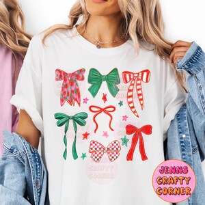 May include: White t-shirt featuring a collection of colorful bow illustrations. The bows are in various designs, including floral, striped, and checkered patterns, with green, red, and pink accents. The text "JENNS CRAFTY CORNER" is in a pink circle.