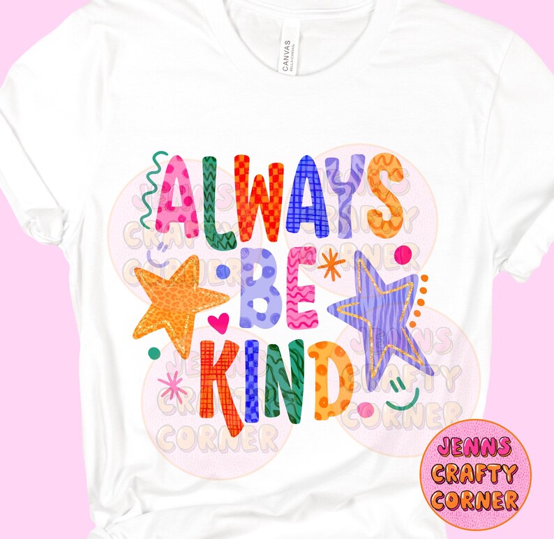 May include: White t-shirt with the words "ALWAYS BE KIND" in colorful, playful lettering. The design includes stars, hearts, and decorative elements. A small logo in a pink circle is in the bottom right corner.