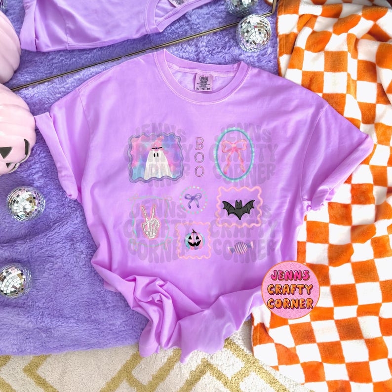 May include: A light purple t-shirt with multiple Halloween-themed graphics, including a ghost, bow, bat, and pumpkin. The text "Jenns Crafty Corner" is printed on the shirt. The shirt is folded, and a patterned blanket is in the background.